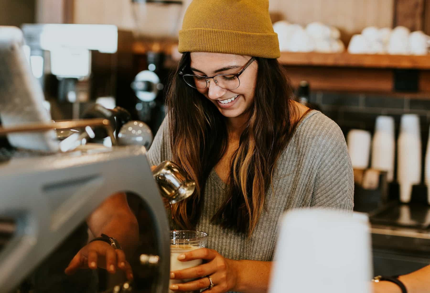 A female barrista with a hat and glasses smiles while making coffee